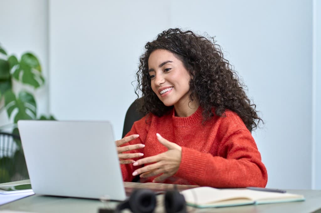 A smiling woman with curly hair in an orange sweater gesturing during a video call on her laptop in a bright, modern office