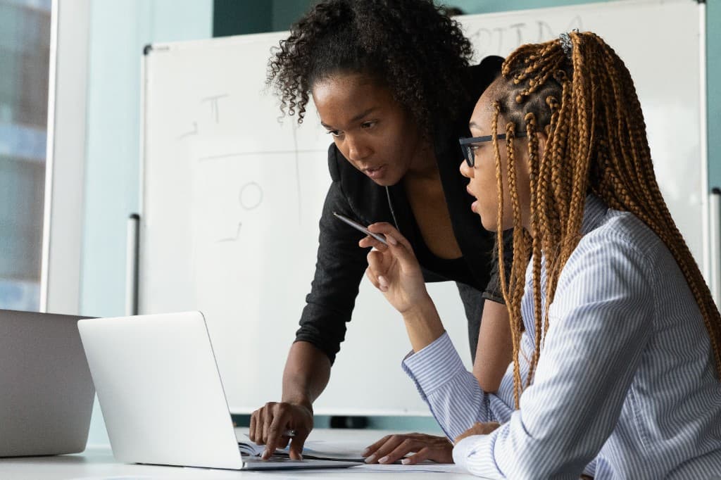 Two Black women in a professional office setting collaborating over a laptop. One woman with braids and glasses is seated, while the other in a dark blazer stands and points at the screen with a pen during a discussion