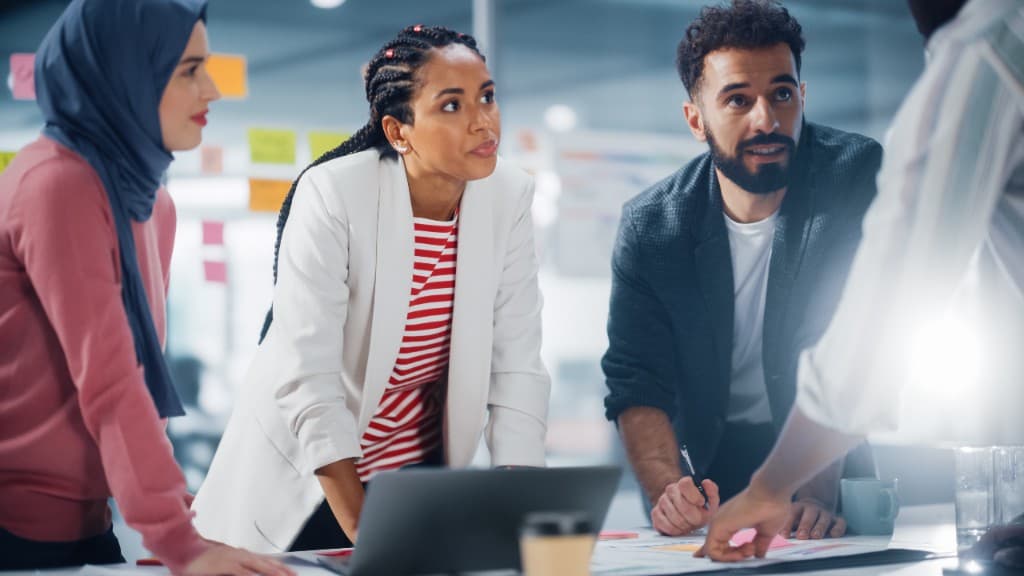 Three diverse colleagues collaborating at a table with a laptop and documents in a bright modern office