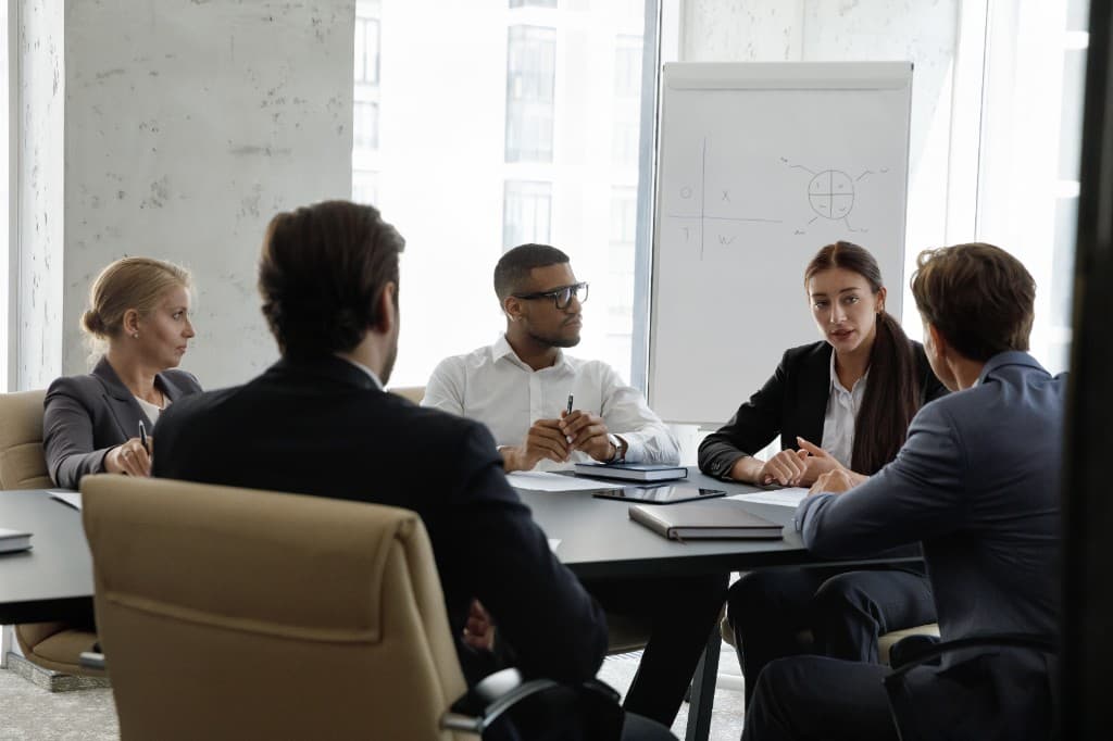 Five professionals in discussion around a conference table in a bright meeting room, with charts on a flip chart in the background