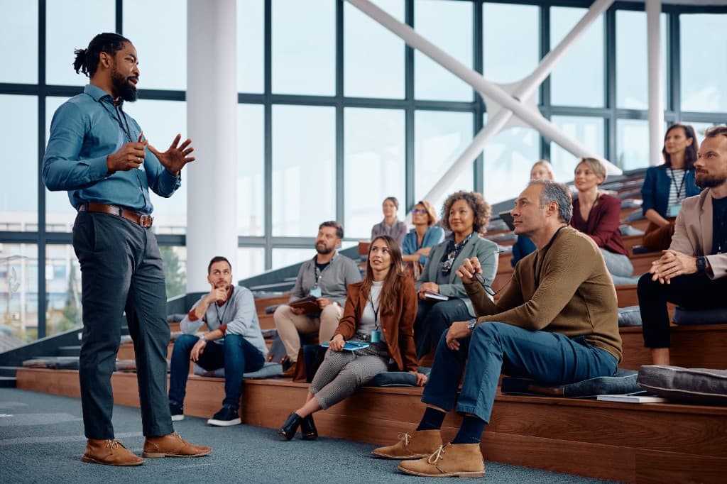 Facilitator presenting to a diverse audience in a bright, modern learning space with tiered seating