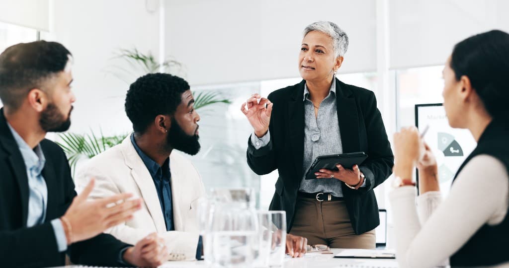 A diverse group of professionals in a modern office meeting, with a woman leader standing and presenting while holding a tablet