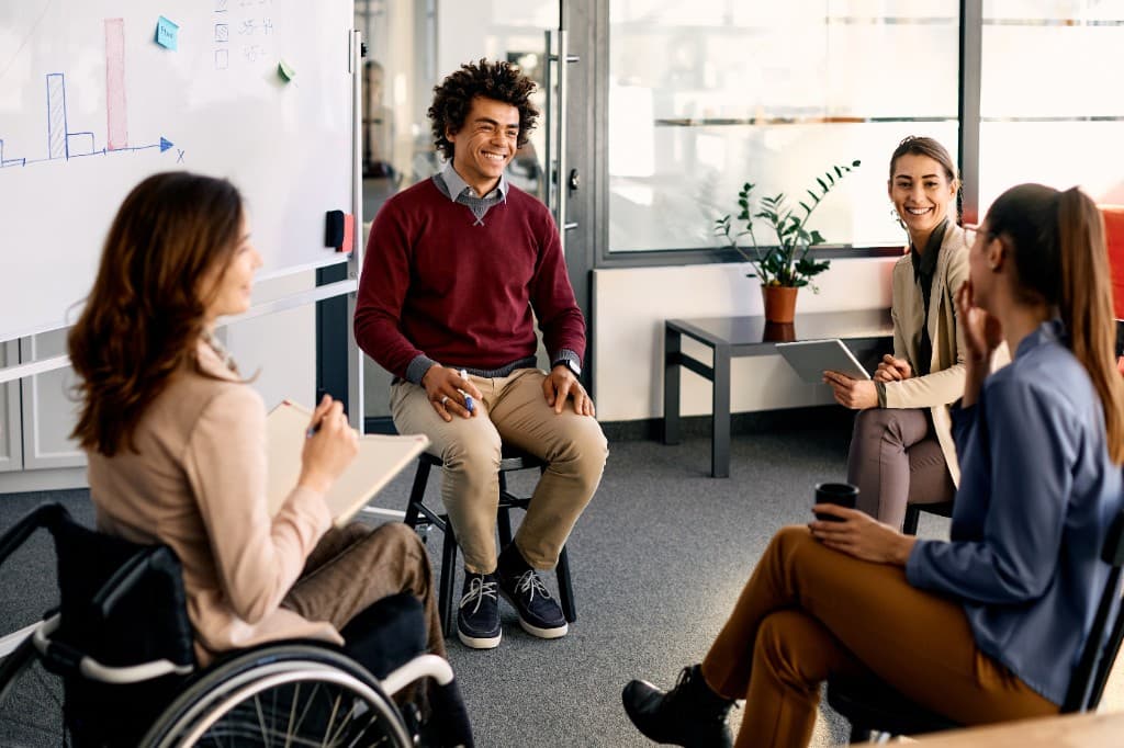 A diverse group of four professionals, including a woman in a wheelchair, having a positive collaborative meeting in a bright, modern office with a whiteboard in the background